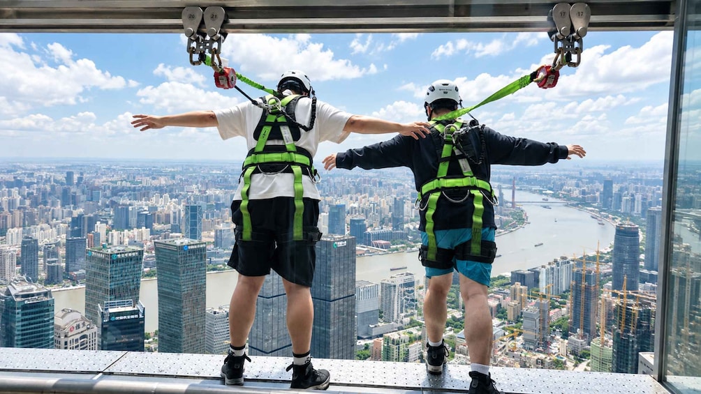 Two people bungee jumping with harnesses and safety gear, overlooking a cityscape with buildings and a river. 3