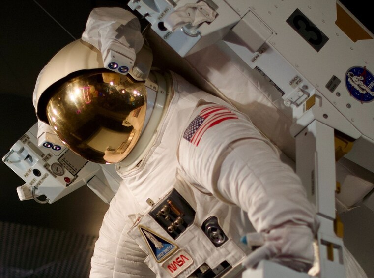 An astronaut in a white spacesuit with an American flag patch, helmet visor, and various equipment on the suit. 5
