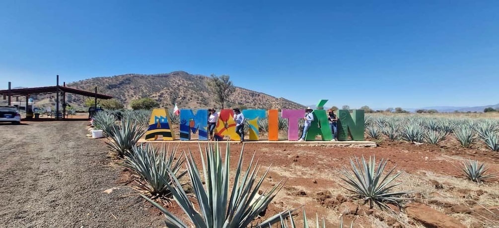 A colorful mural with people standing in front of it, a gas station, and agave plants in a desert landscape. 3
