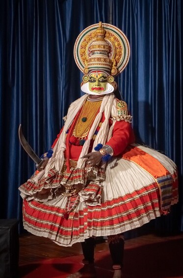A person in traditional costume with a large headdress, holding a sword, and wearing a skirt with red and white stripes. 5
