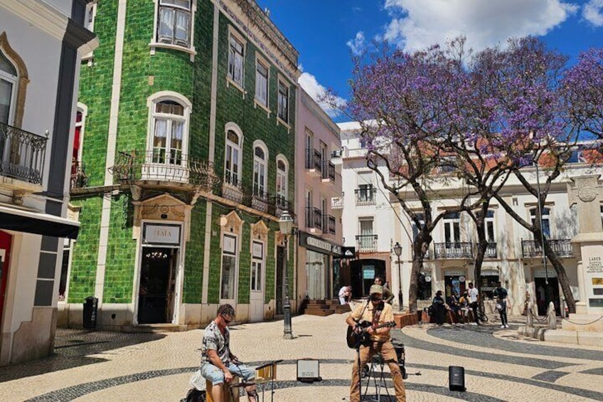 A street with historic buildings, a tree with purple flowers, and a person playing a guitar. 2