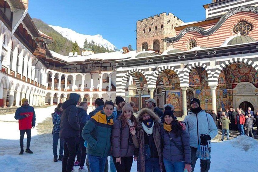 A group of people in a courtyard with snow on the ground, a large building with arches, and a castle-like structure in the background. 3
