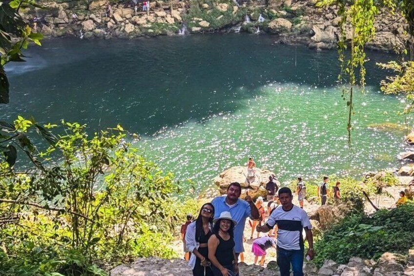 Eine Gruppe von Menschen auf einer felsigen Aussichtsplattform an klarem, türkisfarbenem Wasser mit einem Wasserfall im Hintergrund. 2