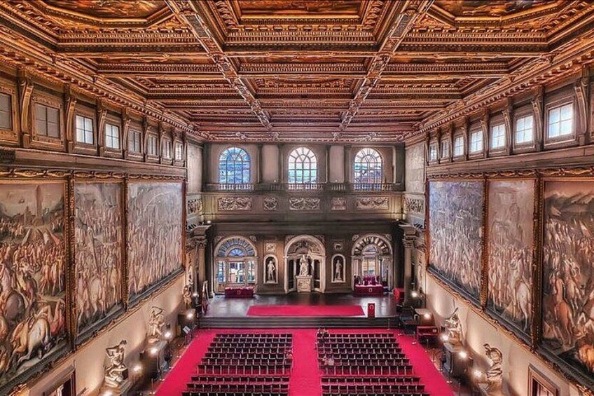 An ornate hall with a red carpet, large paintings, and a central stage. 1