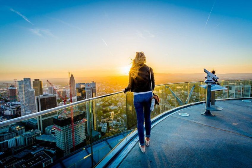 Une personne se tient sur une plateforme d’observation en verre, d’où l’on peut admirer un panorama sur la ville au coucher du soleil. 1