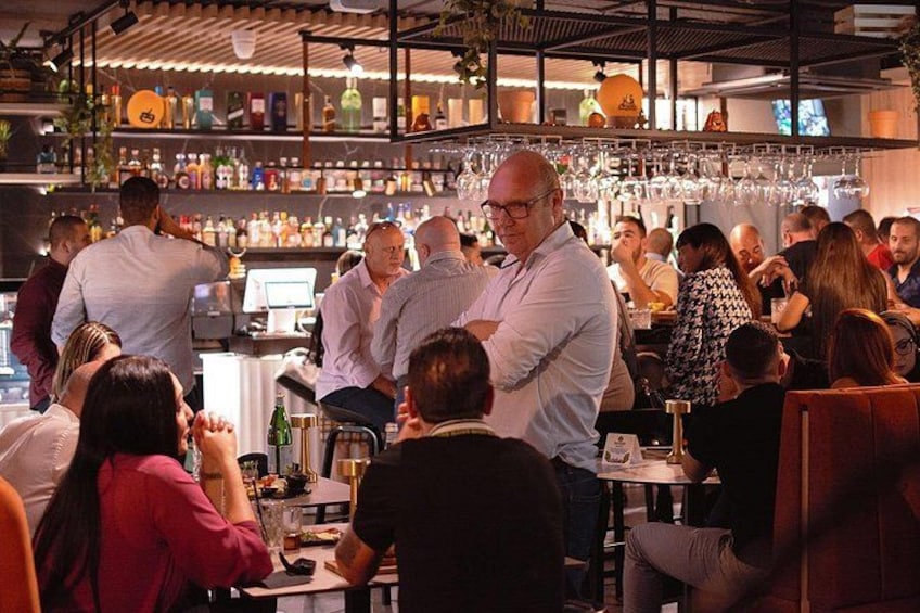 A crowded bar with people sitting and standing, a bartender, and shelves with bottles and glasses. 3