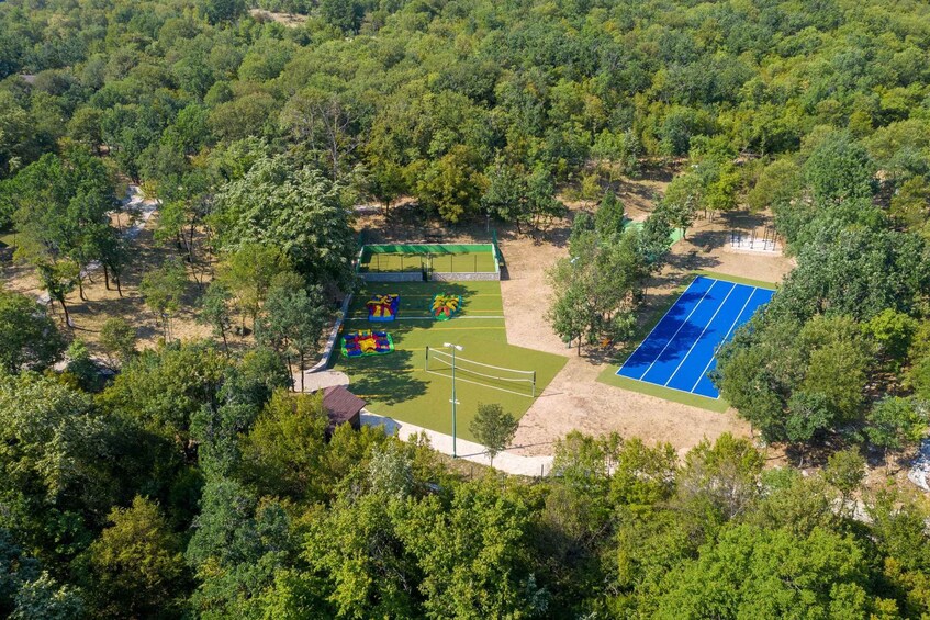 An aerial view of a park with a tennis court, a green sports field, and surrounding trees. 1