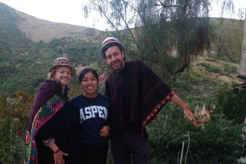 Three people outdoors, one wearing a hat, another in a dark shirt, and the third in a shawl, with a mountainous landscape in the background. 1