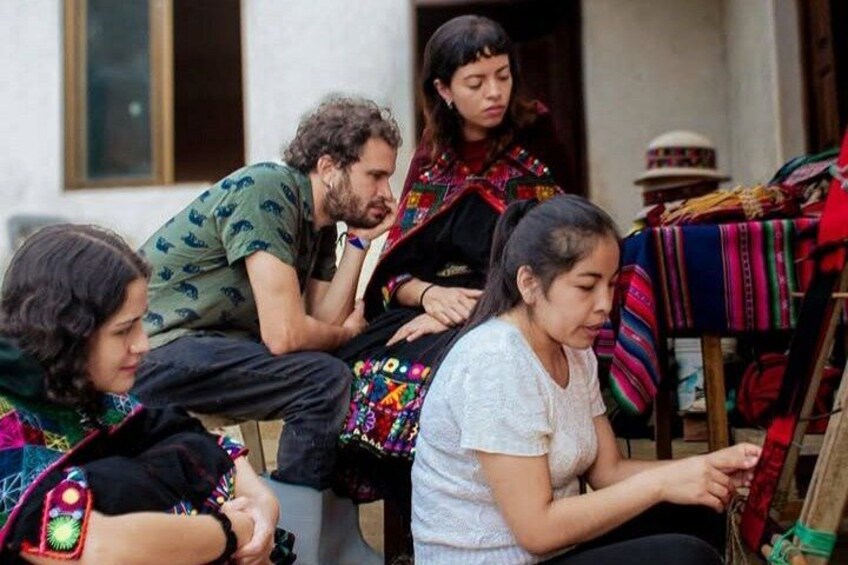 A group of people sitting outdoors, some engaged in conversation, with colorful textiles displayed on a table. 5