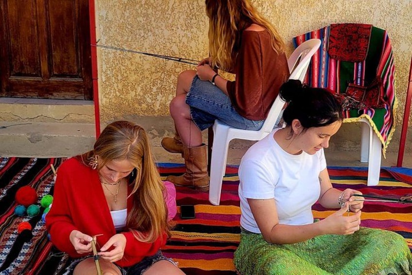 Three people sitting on colorful rugs, one knitting, another holding a phone, and a third in the background. 4