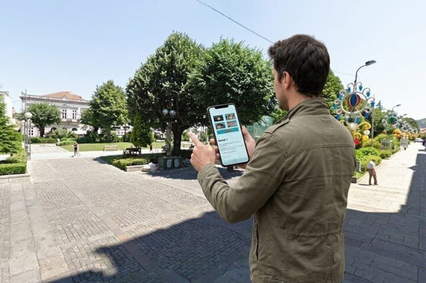 Une personne utilise un téléphone intelligent dans un environnement extérieur, avec des arbres, une allée pavée et un bâtiment en arrière-plan. 2