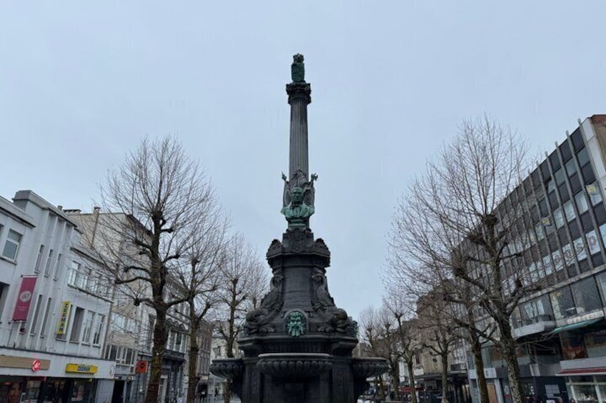 A tall monument with a statue on top, surrounded by leafless trees and modern buildings. 2