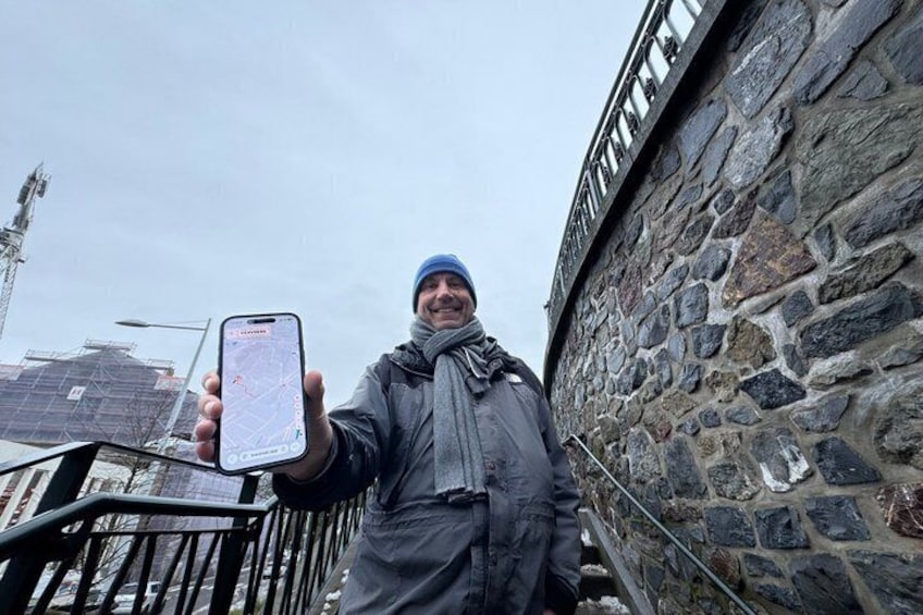 A person holding a smartphone with a map application, standing next to a stone wall with a metal railing. 5