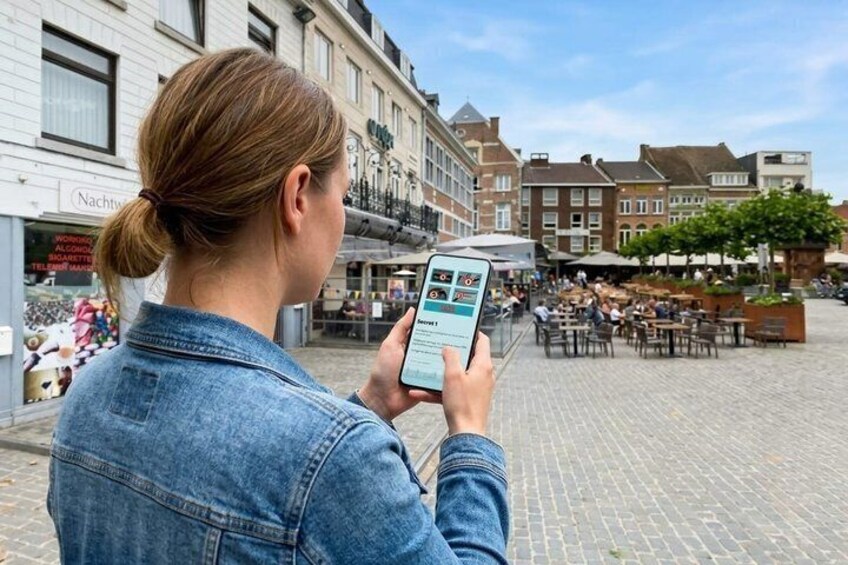 A person holding a smartphone in an outdoor urban setting with cobblestone pavement, historic buildings, and outdoor seating areas. 4