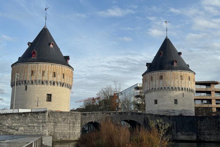 Deux tours historiques surmontées de toits coniques, un pont de pierre et, au loin, des bâtiments modernes. 1