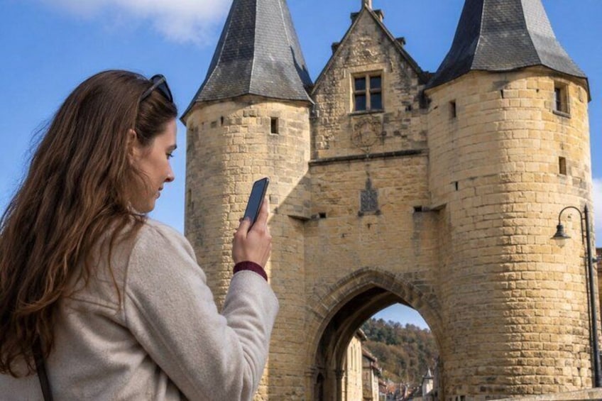 A person taking a photo of a stone tower with a pointed roof and arched entrance. 2