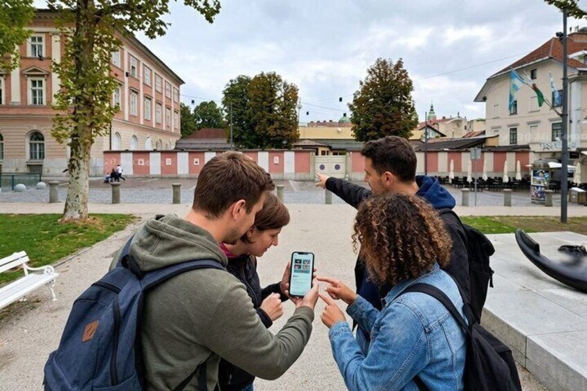 Drie personen maken een foto met een smartphone in een stedelijke omgeving, met gebouwen en een boom op de achtergrond. 3