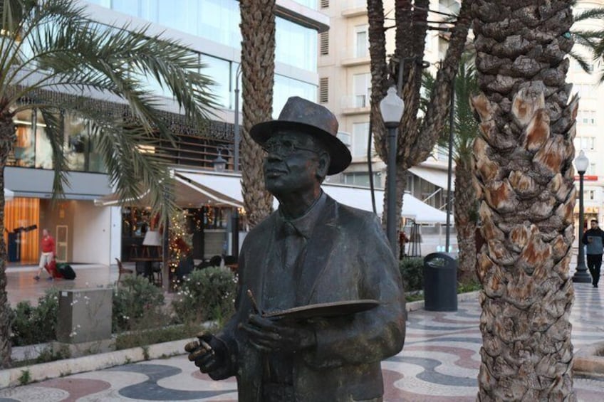 Een standbeeld van een man die een boek vasthoudt, op een plein met palmbomen en moderne gebouwen op de achtergrond. 3