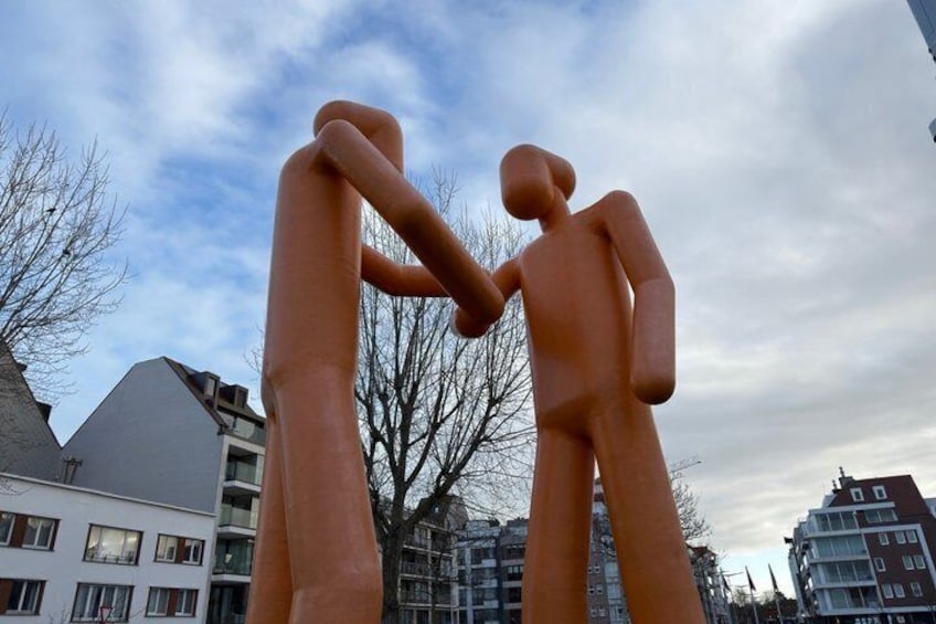 A sculpture of two figures in a heart shape, set against a backdrop of buildings and a cloudy sky. 1