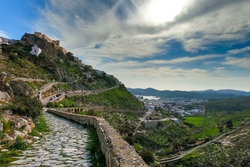 A stone pathway leading to a fortified hilltop structure, a town below, and a valley with greenery. 1