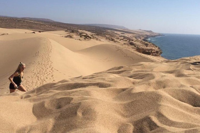 A person walking on sand dunes with a view of the ocean and distant hills. 5