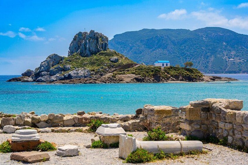 A coastal landscape with ruins, a prominent rock formation, and a small building on a hill. 1