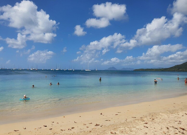 A beach with clear turquoise water, people swimming and sunbathing, and sailboats in the distance. 3