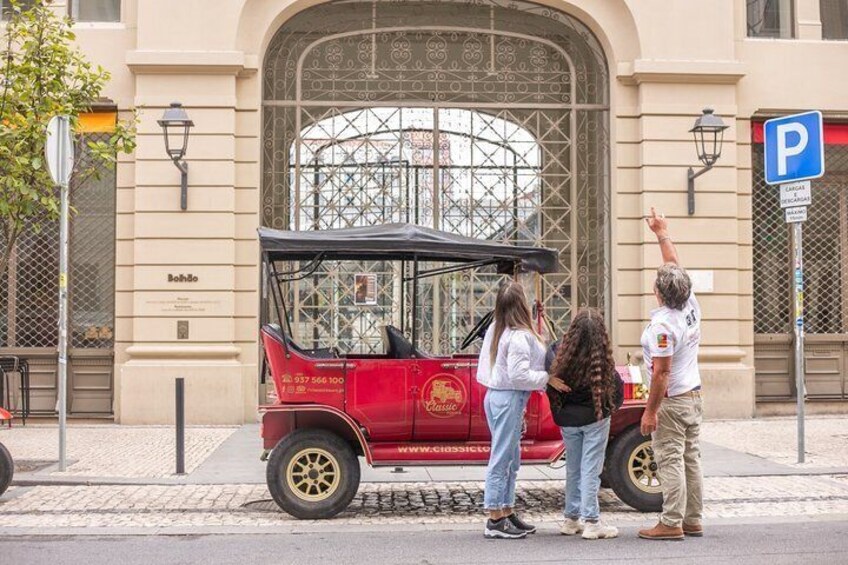 En röd, klassisk bil med svart tak står parkerad på en kullerstensgata. I närheten finns en blå parkeringsskylt och en byggnad med ett stort, välvt fönster och en skylt med texten ”Bohndorf”. 4