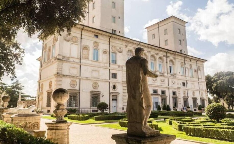 A classical building with a statue in the foreground, surrounded by manicured gardens and a clear sky. 3