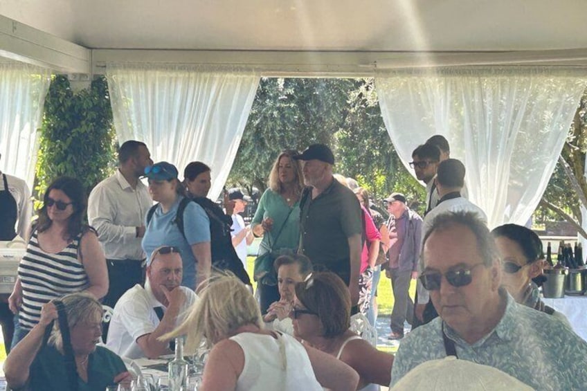 People seated at tables under a tented area with white drapes. 4