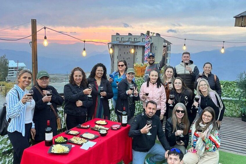 A group of people at an outdoor event with a table of food and drinks, a sunset in the background, and a building in the distance. 2