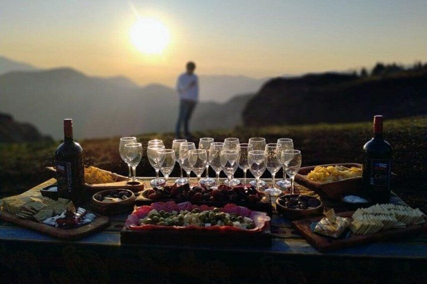 A picnic setup with wine glasses, bottles, and food on a table against a mountainous backdrop during sunset. 1