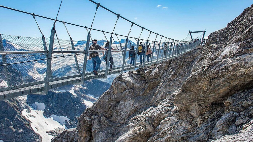 A suspension bridge with people walking across it, surrounded by rocky terrain and snow-capped mountains. 1
