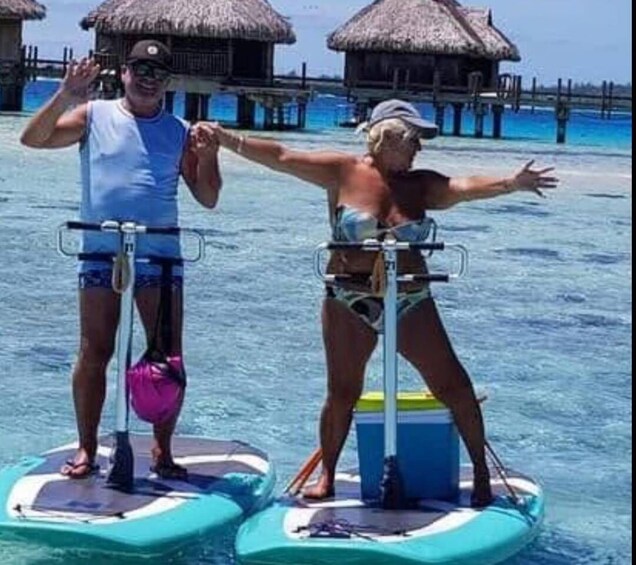 Two people on stand-up paddleboards in a clear blue sea with overwater bungalows in the background. 5