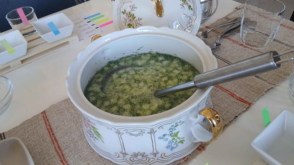 A ceramic bowl with a ladle, filled with a green soup, on a table with a striped cloth. 2