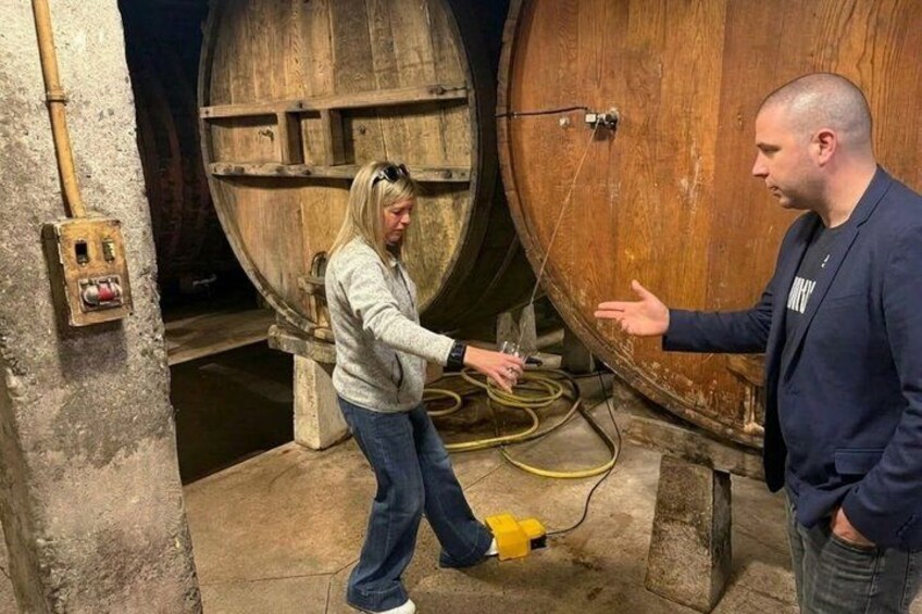 Dos personas interactúan cerca de un barril de madera grande en una bodega. 1