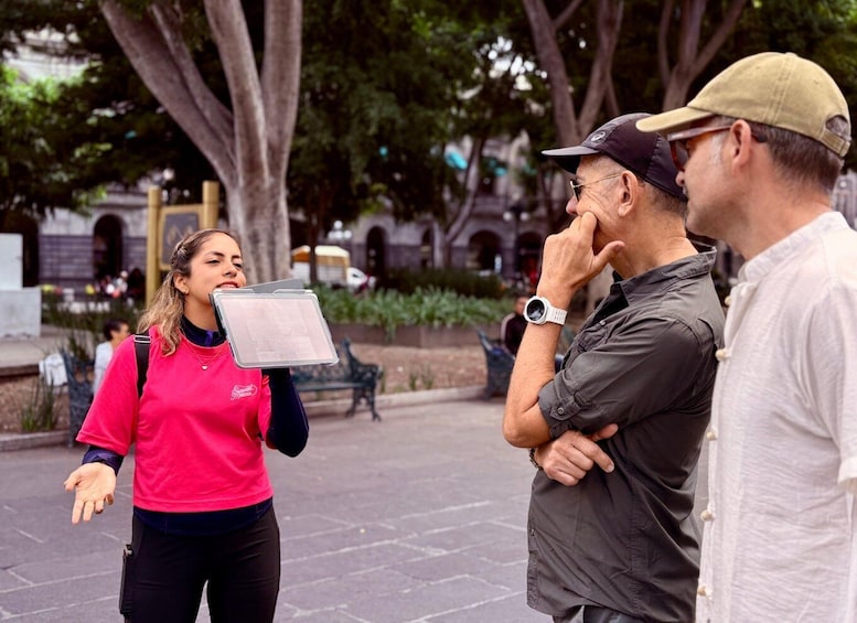 A person in a pink shirt holding a clipboard, standing in a public square with trees and benches. 3