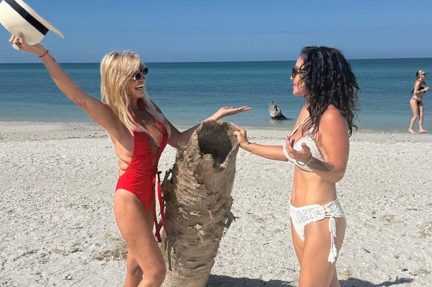 Two people on a beach, one in a red swimsuit and the other in a white bikini, interacting with a large piece of driftwood. 1