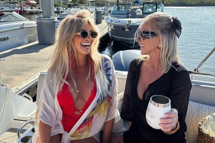 Two people on a boat, one holding a mug, with a marina and boats in the background. 2