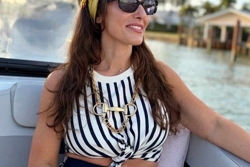 A person wearing a striped top and a gold necklace, seated on a boat with water and buildings in the background. 5
