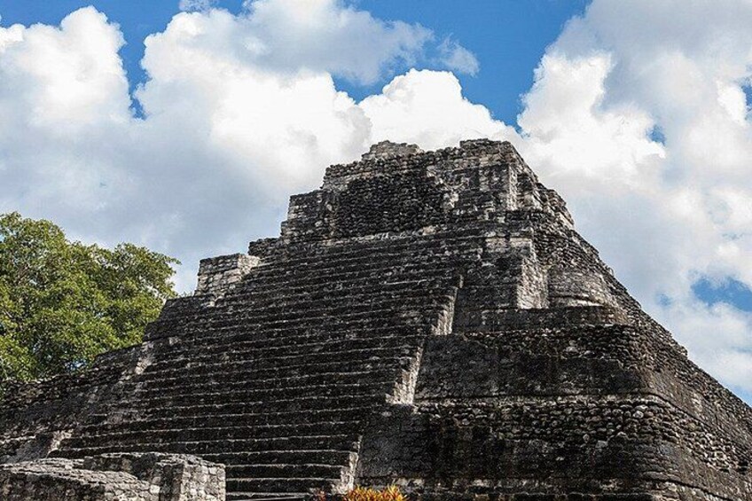 En pyramidliknande, forntida byggnad med trappsteg som leder upp till en platt topp, omgiven av träd och en delvis molnig himmel. 3