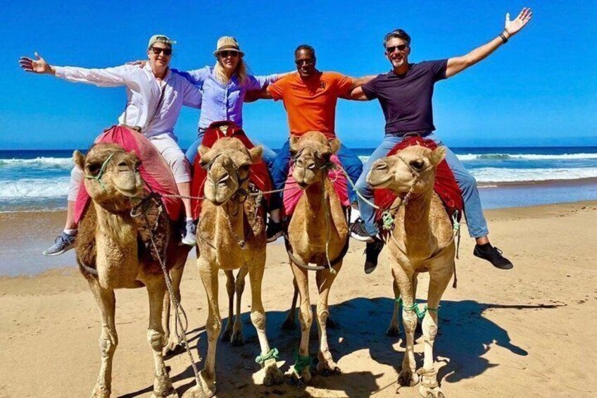Four people riding camels on a beach with the ocean in the background. 1