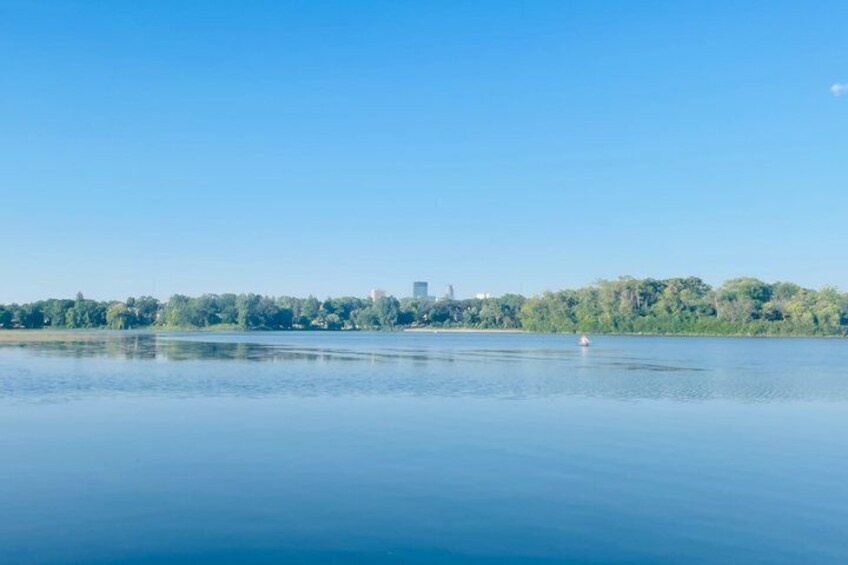 A calm lake with a clear sky, a distant city skyline, and a line of trees along the shore. 2
