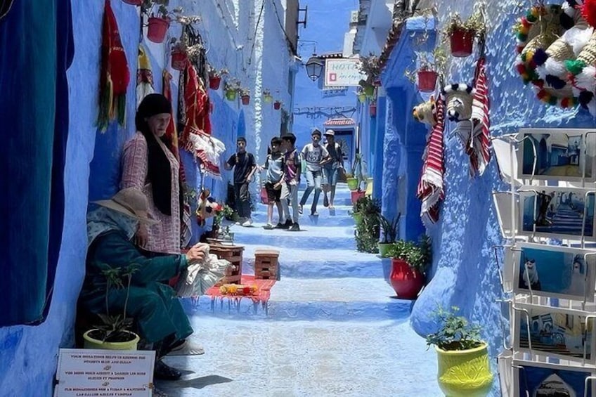 Une rue étroite aux murs bleus, bordée de plantes en pot, où l’on voit des gens qui se promènent. 5