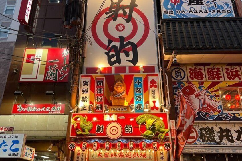 En livlig stadsgata med upplysta skyltar och butiksfönster, inklusive en framträdande röd och vit skylt med japanska tecken. 4