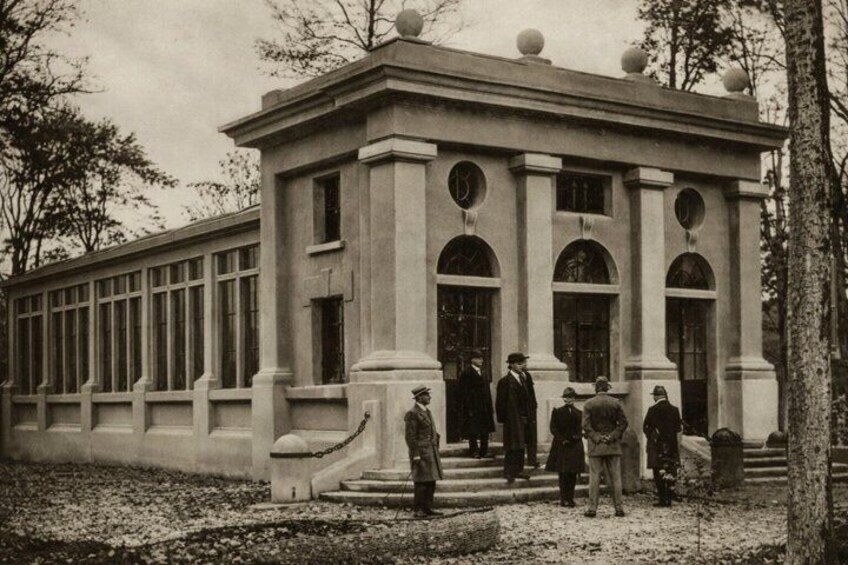 Un bâtiment de style classique, doté de colonnes et de fenêtres en arc de cercle, entouré d’arbres et bordé d’un chemin de gravier. 2