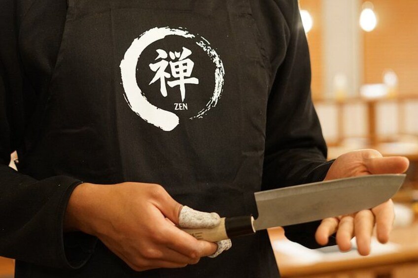 A person wearing a black apron with a white logo and holding a knife. 3
