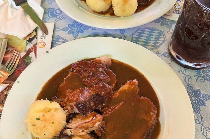 Ein Teller mit Fleisch, Soße, Kartoffelstampf und einem Glas dunkles Getränk auf einem Tisch mit gemustertem Tuch. 2