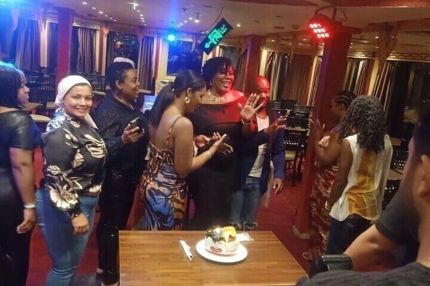 A group of people gathered around a table with a cake, in a room with curtains and a stage in the background. 5