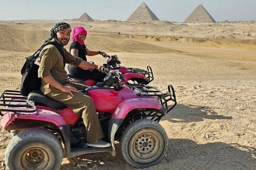 Two people on a pink ATV in a desert with pyramids in the background. 1